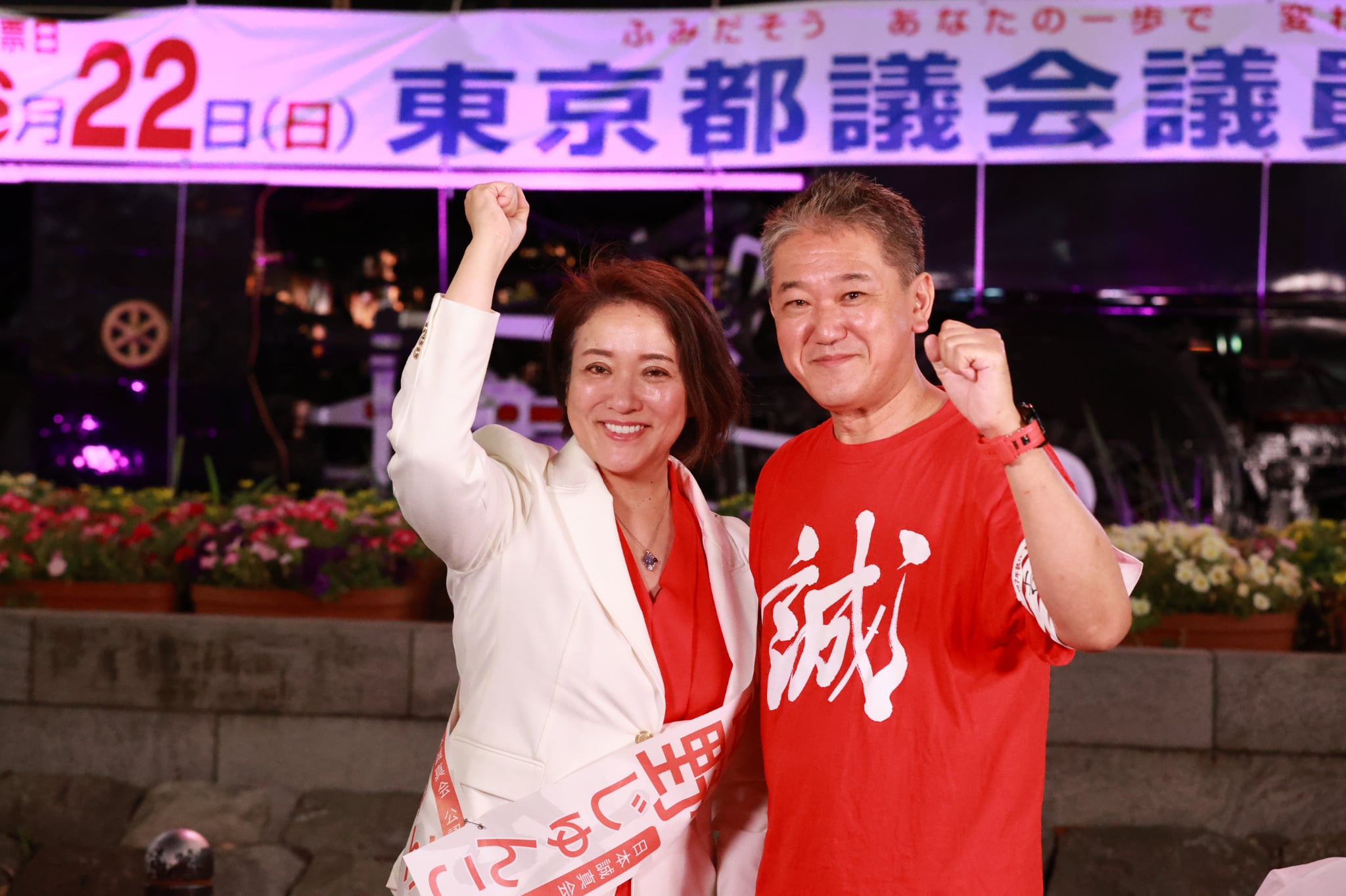 夜、白いジャケットの女性と並び、拳を上げる吉野敏明